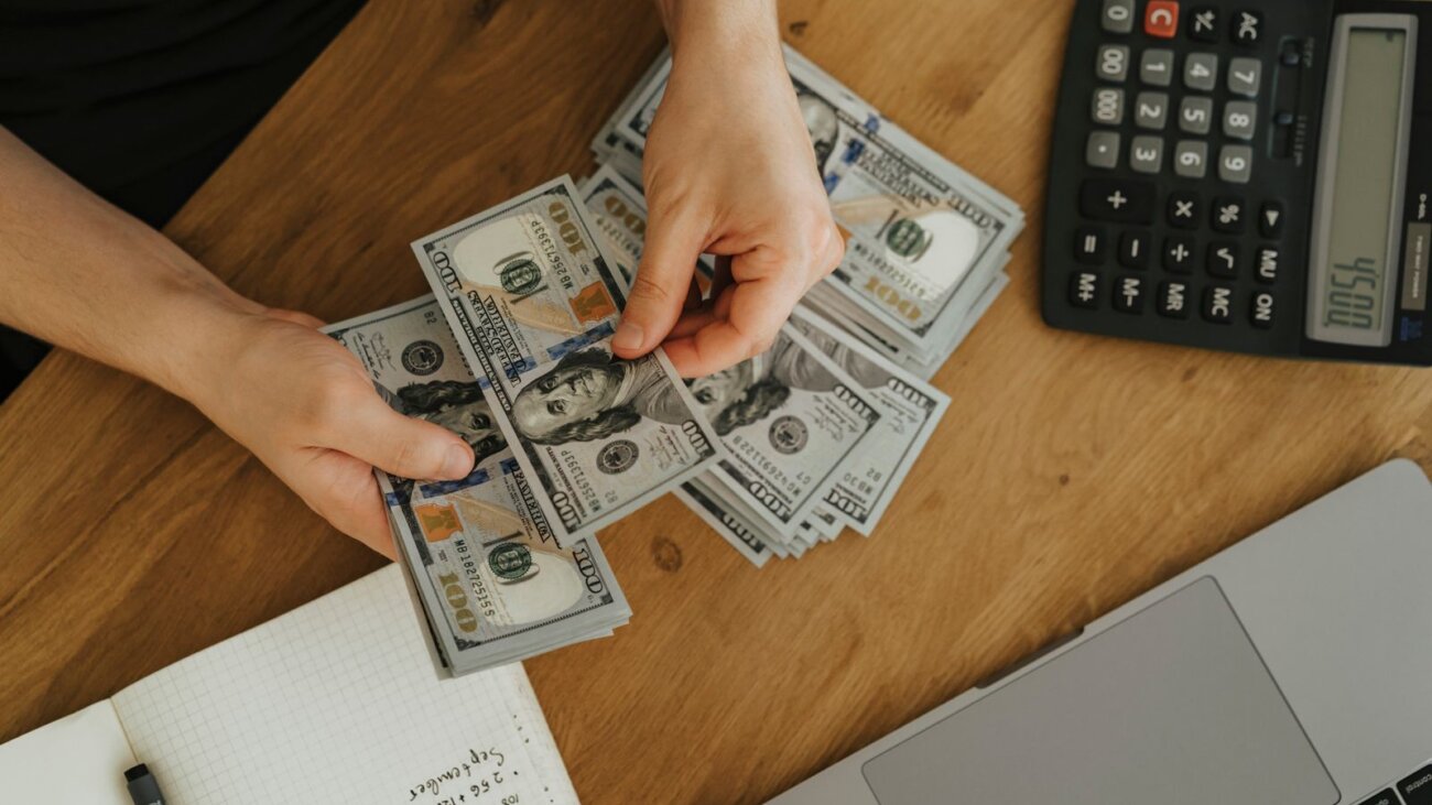 Hands counting dollar bills with a calculator and laptop to budget a software project